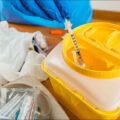 Used syringe being safely disposed of in a yellow sharps container with medical gloves and supplies nearby